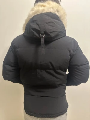 Svart dunjacka med päls från Moose Knuckles - Svart dunjacka från Moose Knuckles med ikonisk metall-logga på ärmen, fluffig avtagbar huva med ljus päls, dragkedja och knappar framtill samt flera fickor. Jackan har en schysst puffer-look och är perfekt för kalla dagar.