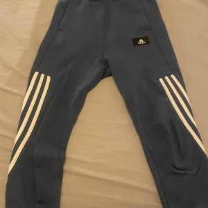 Blå Adidas mjukisbyxor med vita ränder - Snygga blå mjukisbyxor från Adidas med klassiska vita ränder längs benen och Adidas-logga på höften. Byxorna har elastisk midja och är gjorda i mjukt bomullsmaterial, perfekta för chill eller träning. Passformen är normal och benen är raka med ribbade muddar.