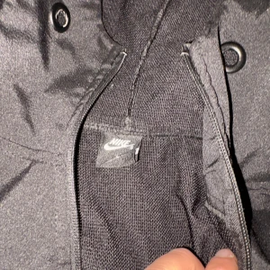 Nike Dri-Fit svart och vit vindjacka - Nike Dri-Fit vindjacka i svart och vitt med halv dragkedja, huva och flera fickor med dragkedja. Jackan har coola kontrastpaneler och logga på bröstet samt ärmen. Perfekt för dig som gillar sportig och clean stil.