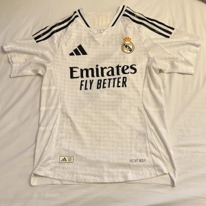 Real Madrid Mbappé #9 fotbollströja - Säljer en officiell Real Madrid fotbollströja från Adidas med Mbappé och nummer 9 på ryggen. Tröjan är vit med svarta detaljer, korta ärmar och har klubbmärket samt sponsortryck på bröstet. Tillverkad i lätt och ventilerande material med HEAT.RDY-teknologi.