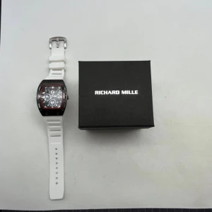 Richard Mille vit sportklocka - Säljer en Richard Mille klocka med vit sportig rem och rektangulärt ur i svart och rött. Urtavlan har flera detaljer och synliga skruvar runt boetten. Klockan levereras med original ask. Perfekt för dig som gillar exklusiv design och coola detaljer.