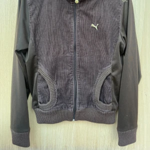 Brun manchester retrojacka från Puma - Snygg brun retro jacka från Puma i manchester med ribbade muddar och hög krage. Jackan har dragkedja framtill och långärmade släta ärmar i matchande mörkbrunt tyg. Puma-logga och extra detalj broderad på axeln i guld.  Storleken är för dam men jackan passar även killar - men då är jackan snarare stlk Small.  Jackan är i bra skick - men är använd samt köpt för ca 20 år sedan...