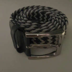Svart och grått flätat bälte - Storlek 115 tror jag.     Snyggt flätat bälte i svart och grått med ett randigt mönster. Bältet har en silvrig metallspänne och detaljer i svart skinn vid ändarna. Perfekt för att ge din outfit en cool touch och passar till jeans eller chinos.