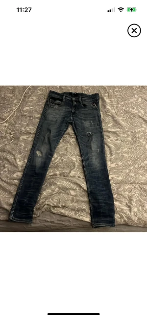 Blå skinny jeans med slitningar - Säljer ett par blå skinny jeans med coola slitningar och tvättade detaljer. Jeansen har fem fickor, normal midja och klassisk knappstängning. Passformen är tight och de sitter snyggt längs benen. Perfekta för dig som gillar en modern streetstil.