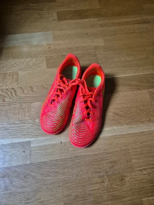 Adidas neonröda fotbollsskor - Säljer ett par Adidas fotbollsskor i neonrött med coola mönster i gult på ovansidan. Skorna har snörning och platt sula, tillverkade i syntetmaterial för lätt känsla på planen. Perfekta för dig som vill sticka ut med färg och stil under matchen.