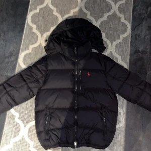 Svart pufferjacka från Polo Ralph Lauren - Svart pufferjacka från Polo Ralph Lauren med röd broderad logga på bröstet. Jackan har huva, två sidofickor med dragkedja och en klassisk quiltad design. Perfekt för kalla dagar och har en snygg, sportig look.