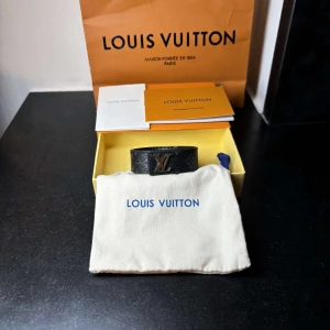 Louis Vuitton svart armband med LV-logo - Snyggt svart armband från Louis Vuitton i äkta skinn med klassisk LV-logo i silver på framsidan. Armbandet har diskret monogrammönster och justerbar passform. Perfekt accessoar för dig som gillar exklusiva detaljer och stilren design.