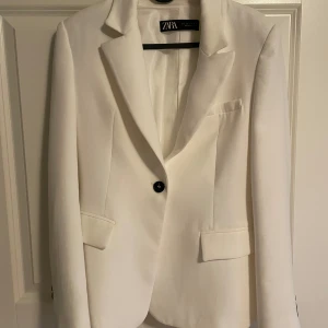 Vit kavaj från Zara, storlek S - Snygg vit kavaj från Zara i klassisk modell med smal passform. Kavajen har svarta knappar framtill och vid ärmslut, samt fickor med lock. Materialet är mjukt och har en lätt glans. 
