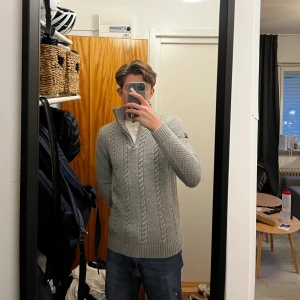 Grå stickad half zip-tröja Superdry - Säljer en grå stickad tröja från Superdry med kabelstickat mönster och hög krage med half zip. Tröjan har långa ärmar, ribbade muddar och en snygg Superdry-patch på ärmen. Perfekt för kyliga dagar och riktigt skön att ha på sig.                      Nypris: 1000kr