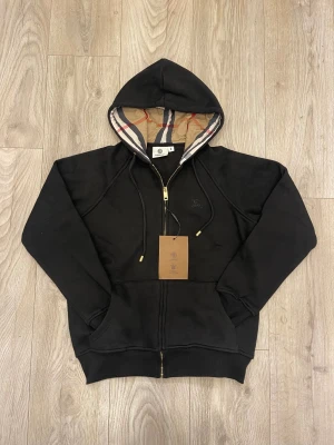 Svart hoodiejacka från Burberry - Svart hoodiejacka från Burberry med klassiskt rutigt foder i huvan, guldfärgad dragkedja och snörning. Jackan har broderad logga på bröstet och ribbade muddar. Perfekt för dig som vill ha en stilren och lyxig look.