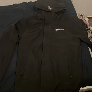 Svart regnjacka från Tenson med huva - Svart regnjacka från Tenson med justerbar huva och dragkedja framtill. Jackan är tillverkad i syntetmaterial med MPC-beläggning som gör den både vattentät och vindtät. Perfekt för blöta och blåsiga dagar, med stilren design och logga på bröstet.