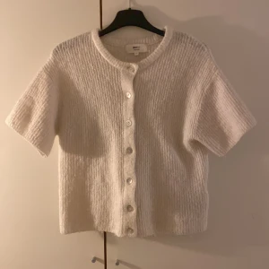 Vit stickad kofta från Gina Tricot - Mysig vit stickad kofta från Gina Tricot i ullblandning. Koftan har korta ärmar, rund hals och knappar framtill. Perfekt för lager-på-lager och har en ribbad struktur som ger en chill vibe.