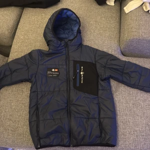 Blå Sail Racing pufferjacka med huva - Snygg mörkblå pufferjacka från Sail Racing med huva och coola patchdetaljer på bröstet. Jackan har en svart bröstficka med dragkedja och Sail Racing-logga, samt quiltad design. Perfekt för kalla dagar och har en sportig vibe.