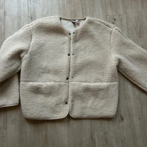 Beige teddyjacka från Uniqlo - Supermjuk och oversized teddyjacka från H&M i beige. Jackan har rund hals, tryckknappar framtill och två stora fickor. Perfekt för lager-på-lager och chill vibes. Fleece-liknande material som är både varmt och skönt.