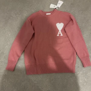 Rosa stickad tröja  - Snygg rosa stickad tröja från Ami med rund hals och ribbade muddar. På bröstet finns ett vitt hjärta med bokstaven A, vilket ger en unik och trendig vibe. Perfekt för dig som gillar stilrena detaljer och vill ha något som sticker ut.