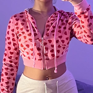 Croppad rosa hoodie med hjärtan - Säljer en croppad hoodie i ljusrosa med röda hjärtan över hela tröjan. Tröjan har dragkedja framtill, snörning vid huvan och är tillverkad i mjuk velour. Långärmad och med bred mudd nedtill. Perfekt för dig som gillar gulliga detaljer.