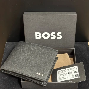 Hugo Boss plånbok i grained läder, helt ny, oanvänd - Helt ny och oanvänd plånbok från Hugo Boss i grained (kornigt) läder. Stilren svart design med Hugo Boss-logo. Plånboken har flera kortfack, fack för sedlar och ett myntfack. Följer med originalförpackning. Nypris för liknande modeller ligger runt 1 000–1 300 kr.   Denna säljs för ett mycket konkurrenskraftigt pris – perfekt för dig som vill ha premiumkvalitet till ett bra värde.
