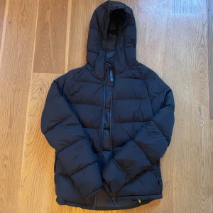 Svart pufferjacka med huva - Svart pufferjacka från Weekday med huva och dragkedja framtill. Jackan har en quiltad design och är tillverkad i syntetmaterial. Perfekt för kalla dagar med sin tjocka och fluffiga look. Passformen är normal och jackan har långa ärmar. Passar som S