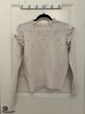 Vit stickad tröja med pärlor och volang - Supersöt vit stickad tröja med långärmad design och volangdetaljer vid axlarna. Tröjan har ett snyggt diamantmönster framtill och är dekorerad med pärlor och små glittriga stenar. Ribbad kant vid hals, ärmslut och nederkant. Perfekt för dig som gillar detaljer!