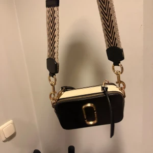 Marc Jacobs Snapshot  - Säljer en Marc Jacobs Snapshot axelväska i svart och vitt med guldiga metalldetaljer och bred, justerbar axelrem.  (Bandet som ingår är originalet till den svartvita väskan vilket är den på 3 bilden) de 2 andra banden på bild 1,2 går att köpa för 700kr/st nypris 1100-1200kr