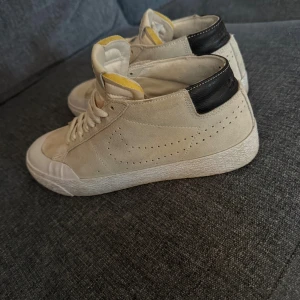 Nike SB Blazer Mid i beige mocka - Nike SB Blazer Mid sneakers i beige mocka med svart häl och vit sula. Skorna har snörning, perforerat swoosh-mönster på sidan och gul detalj på plösen. Klassisk rund tå och platt sula, perfekt för dig som gillar skate och streetstyle. Storlek 41
