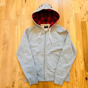 Grå hoodie från Stüssy med röd rutig huva - Grå zip-hoodie från Stüssy med röd- och svart-rutig insida i huvan och broderad Stüssy-logga på huvan. Tröjan har dragsko i huvan, två fickor fram och är i mjuk bomullsmix. Perfekt för en chill streetwear-look.