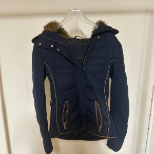Marinblå pufferjacka från Zara Basic - Snygg marinblå pufferjacka från Zara Basic med fuskpäls på luvan och guldiga detaljer. Jackan har dragkedja, knappar och två fickor med zip framtill. Beige piping längs sidorna ger en schysst kontrast. Perfekt för kalla dagar!
