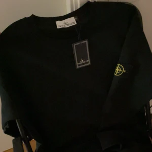 Svart sweatshirt från Stone Island - Svart sweatshirt från Stone Island med klassisk rund hals och ikonisk logotyp-patch på vänster ärm. Tröjan har ribbade muddar och är tillverkad i mjuk bomull. Perfekt för en clean och stilren streetwear-look.