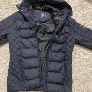 Svart pufferjacka från Vinson Polo Club - Svart pufferjacka med huva från Vinson Polo Club. Jackan har quiltade partier på sidorna och ärmarna, dragkedja framtill och två sidofickor med dragkedja. Märkespatch på vänster ärm och snygga detaljer vid kragen. Perfekt för kalla dagar. För ungdomar 