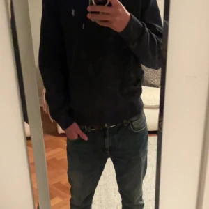 Navy blå hoodie från Polo Ralph Lauren - Svart hoodie från Polo Ralph Lauren med dragkedja och huva med snörning. Klassisk logga broderad på bröstet och två fickor framtill. Skön passform och mjukt bomullsmaterial, perfekt för chill dagar. Snygg och enkel design som funkar till jeans eller joggers.