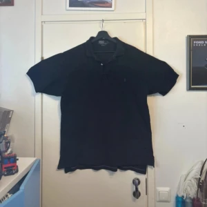 Svart pikétröja från Polo Ralph Lauren - Svart pikétröja från Polo Ralph Lauren i 100% bomull. Klassisk design med krage, korta ärmar och två knappar framtill. Diskret broderad logga på bröstet. Perfekt för en clean och stilren look.