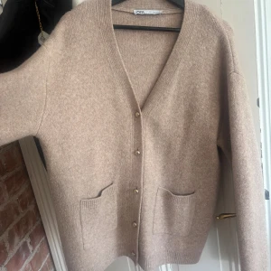 Beige kofta från Zara med fickor - Mysig beige kofta från Zara i storlek S. Koftan har v-ringning, guldfärgade knappar och två praktiska fickor framtill. Materialet är stickat och känns mjukt mot huden. Perfekt för lager-på-lager och chill dagar.