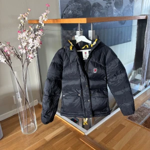 Svart dunjacka Fjällräven XS - Absolut i nyskick. Använd ett fåtal gånger då den var något för liten för min del. Köpt vintern 2025. Svart dunjacka från Fjällräven Expedition Series med huva och gula detaljer vid snörningen. Jackan har två stora fickor framtill med knappar och dragkedja, samt klassisk röd Fjällräven-logga på bröstet. Perfekt för kalla dagar och riktigt snygg passform.❤️