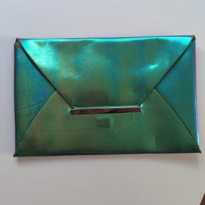 Grön metallic kuvertväska - Snygg kuvertväska i metallicgrönt med skimrande yta och guldsilvrigt innerfoder. Väskan har en stilren, rektangulär form och stängs på en metall-detalj framtill. Perfekt för dig som vill sticka ut med en färgstark accessoar. Mått: längd 28 cm bred 18 cm