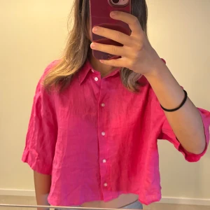 Rosa croppad linneskjorta från H&M - Säljer en snygg croppad skjorta i stark rosa från H&M. Skjortan är tillverkad i linne, har knappar framtill och avslappnad passform med trekvartsärmar. Perfekt för dig som gillar färg och vill ha en luftig vibe.