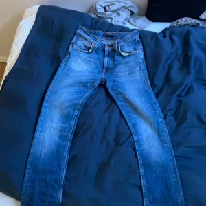 Blå jeans från Nudie Jeans W28 L32 - Snygga blå jeans från Nudie Jeans med klassisk femficksdesign och orangea sömmar. Jeansen har en rak passform och är tillverkade i slitstarkt denim. Perfekta för dig som gillar en tidlös och stilren look.