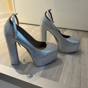 Glitter-silvriga platåpumps  - Säljer ett par silvriga pumps från Call It Spring med hög klack och platåsula. Skorna är täckta av glittrande stenar och har rund tå samt justerbar rem runt vristen. Veganskt material och riktigt statement-skor för dig som vill sticka ut. Skorna är bara använda en gång. 