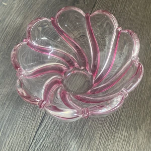 Söt liten skål - Bilderna visar en dekorativ glasskål med rosa detaljer och böljande mönster. Skålen är genomskinlig med rosa ränder och har en unik, blomliknande form. Inga synliga sprickor eller större skador, endast mindre tecken på normalt bruk. Perfekt som inredningsdetalj eller serveringsskål.