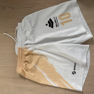 Vita och beige sportshorts BallersApp - Säljer ett par vita sportshorts från Spized med beige detalj och tryck från BallersApp samt siffran 10. Shortsen har elastisk midja med snörning och är gjorda i 100% polyester, perfekta för fotboll eller träning.