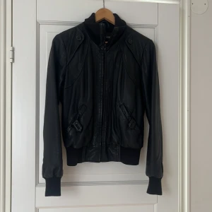Svart skinnjacka i bombermodell M - Svart skinnjacka i bomberstil med dragkedja framtill, ribbade muddar och hög krage. Jackan har coola detaljer som knappar vid axlarna och snedställda fickor. Tillverkad i äkta läder och passar perfekt för dig som vill ha en edgy vibe.