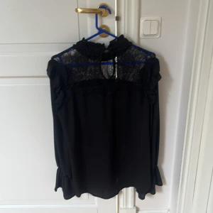 Svart blus med spets från Unika Paris - Supersnygg svart blus från Unika Paris med transparent spets upptill, volangdetaljer på axlarna och vid ärmslut. Hög krage med rynk och knapp i nacken. Perfekt för dig som gillar en elegant och trendig look.