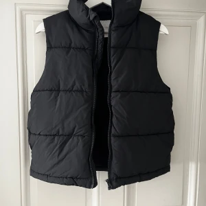 Svart dunväst från Gina Tricot - Svart quiltad dunväst från Gina Tricot med hög krage och dragkedja framtill. Västen har en klassisk, enkel design och är perfekt att slänga över en hoodie eller tröja för extra värme. Passar dig som gillar en stilren look.