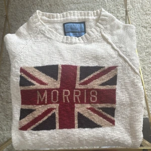 Vit stickad tröja Morris med flagga - Vit stickad tröja från Morris med Union Jack-flagga och texten 'Morris' framtill. Tröjan har rund halsringning och långa ärmar. Snygg kontrast mellan rött, mörkblått och beige i flaggmotivet. Perfekt för dig som gillar klassisk brittisk stil.