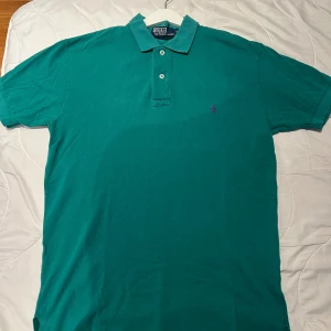 Polo Ralph Lauren  - Klassisk grön pikétröja från Polo Ralph Lauren i storlek S. Tröjan har korta ärmar, krage med två knappar och lila broderad logga på bröstet. Tillverkad i bomull för en skön och luftig känsla. Perfekt för en avslappnad och stilren look. Hör av dig vid fler frågor 