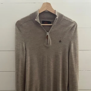 Beige merinoulltröja från Morris - Snygg beige stickad tröja från Morris i 100% merinoull. Tröjan har en ribbad krage med half zip-dragkedja och ett litet broderat emblem på bröstet. Perfekt för dig som gillar stilrena och klassiska plagg med en modern touch.