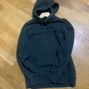 Mörkgrön hoodie från Peak Performance - Säljer en mörkgrön hoodie från Peak Performance med broderad logga på bröstet. Tröjan har huva med snörning, känguruficka och ribbade muddar. Perfekt för chill dagar eller när du vill ha en clean och enkel stil.