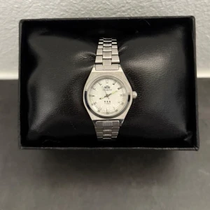 Silverfärgad Orient armbandsur - Stilren klocka från Orient med silverfärgat armband i metall och vit urtavla. Klockan har klassisk rund form, tydliga indexmarkeringar och tre visare. Länken är robust och ger en tidlös känsla. Perfekt accessoar för dig som gillar minimalistisk design.