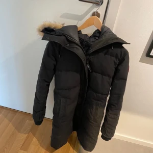 Svart lång dunjacka Canada Goose - Säljer en svart lång dunjacka från Canada Goose med huva och beige pälsdetalj. Jackan har klassisk patch på ärmen, dragkedja och stora fickor framtill. Perfekt för kalla vinterdagar och riktigt snygg passform.