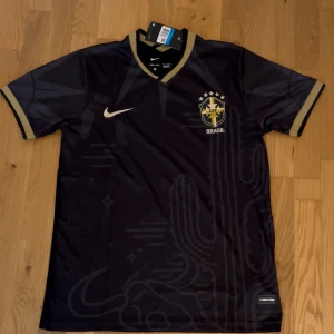 Nike Brasil svart fotbollströja M - Snygg svart Brasilien fotbollströja från Nike med guldiga detaljer och tryckt klubbmärke på bröstet. Tröjan har korta ärmar, V-ringad krage och diskreta grafiska mönster i tyget. Tillverkad i lätt och ventilerande Dri-FIT material som passar perfekt till träning eller match.