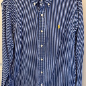 Blåvit randig skjorta från Ralph Lauren - Säljer en klassisk blå och vit randig skjorta från Ralph Lauren med broderad gul logga på bröstet. Skjortan har lång ärm, knappar hela vägen och button-down krage. Tillverkad i mjuk bomull och har en clean, tidlös look.